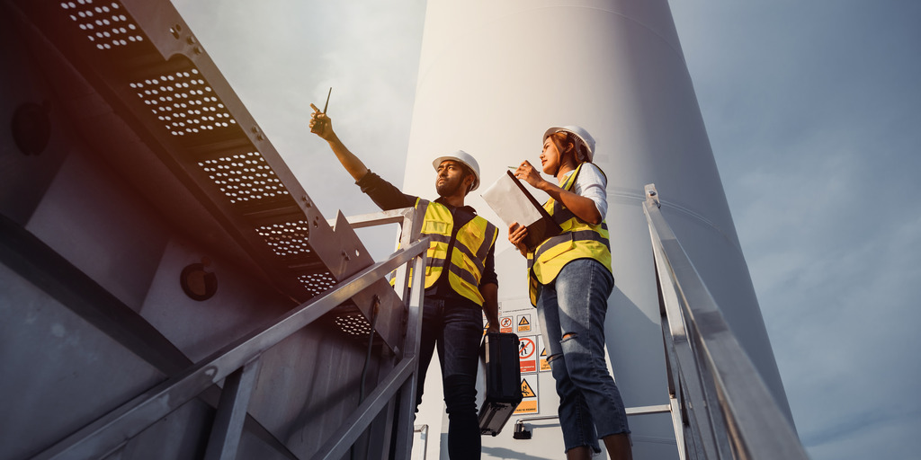 Eine Ingenieurin und ein Ingenieur stehen am Fuß einer Windkraftanlage und blicken in die Ferne.