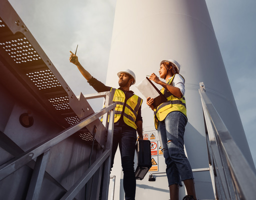 Eine Ingenieurin und ein Ingenieur stehen am Fuß einer Windkraftanlage und blicken in die Ferne.