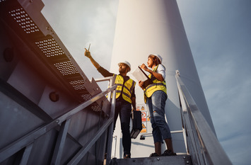 Eine Ingenieurin und ein Ingenieur stehen am Fu&szlig; einer Windkraftanlage und blicken in die Ferne.