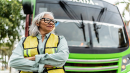 Eine grauhaarige Frau steht als Busfahrerin mit Brille und Warnweste bekleidet vor einem Bus.