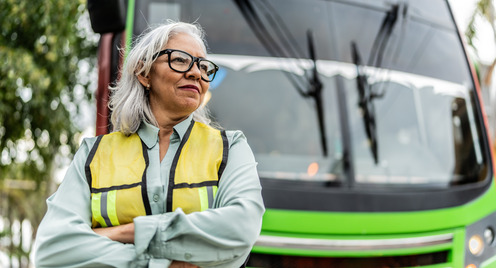 Eine grauhaarige Frau steht als Busfahrerin mit Brille und Warnweste bekleidet vor einem Bus.