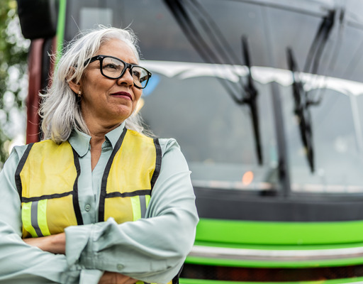 Eine grauhaarige Frau steht als Busfahrerin mit Warnweste bekleidet vor einem Bus.