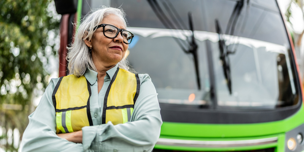 Eine grauhaarige Frau steht als Busfahrerin mit Brille und Warnweste bekleidet vor einem Bus.