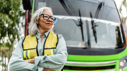 Eine grauhaarige Frau steht als Busfahrerin mit Brille und Warnweste bekleidet vor einem Bus.