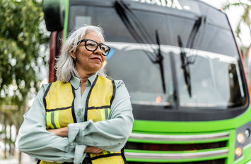 Frau mit gelber Warnweste und verschr&auml;nkten Armen steht vor einem Bus