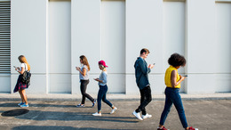 Mehrere Personen laufen auf einem Platz vor einer weißen Wand in entgegengesetzte Richtungen und schauen dabei auf ihr Handy.
