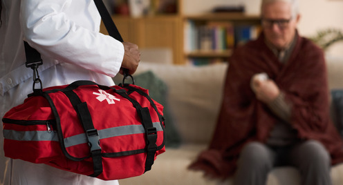 Arstz mit Arzttasche steht vor einem älteren Herren, der sitzend seinen Arm hält