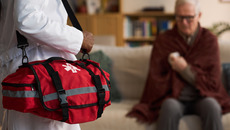 Caucasian senior man sitting on sofa holding arm while Black woman paramedic arriving to assist in foreground carrying red medical bag, home healthcare emergency concept(© everypixel)