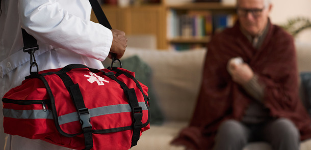 Caucasian senior man sitting on sofa holding arm while Black woman paramedic arriving to assist in foreground carrying red medical bag, home healthcare emergency concept(© everypixel)