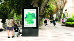 Eine mit Bäumen gesäumte Promenade ist zu sehen. Im Vordergrund steht eine digitale Infotafel mit einer Deutschlandkarte. Auf der Promenade gehen Personen und neben der Infotafal stehen Personen. 