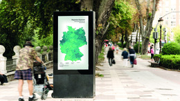 Eine mit Bäumen gesäumte Promenade ist zu sehen. Im Vordergrund steht eine digitale Infotafel mit einer Deutschlandkarte. Auf der Promenade gehen Personen und neben der Infotafal stehen Personen. 