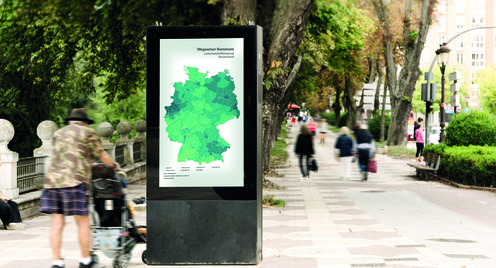Eine mit Bäumen gesäumte Promenade ist zu sehen. Im Vordergrund steht eine digitale Infotafel mit einer Deutschlandkarte. Auf der Promenade gehen Personen und neben der Infotafal stehen Personen. 