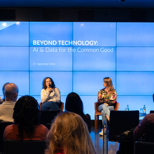 Vier Personen sitzen auf Stühlen vor einer großen Bildschirmwand mit dem Text 'BEYOND TECHNOLOGY: AI & Data for the Common Good 17 September 2026' und sprechen vor einem Publikum