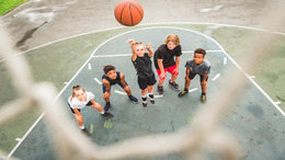 Fünf Kinder stehen auf einem Basketballfeld vor dem Korb. Eines der Kinder wirft den Ball in Richtung Korb.