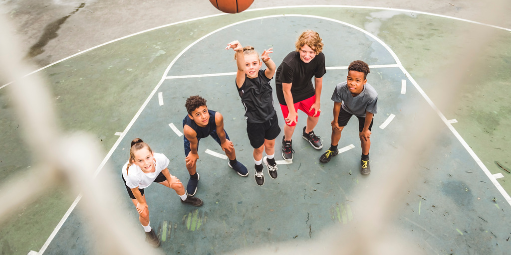 Fünf Kinder stehen auf einem Basketballfeld vor dem Korb. Eines der Kinder wirft den Ball in Richtung Korb.