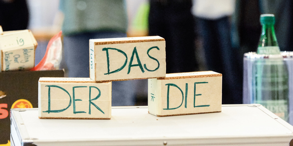 m Vordergrund stehen Holzklötze mit den deutschen Artikeln „der“, „das“ und „die“; im unscharfen Hintergrund sind mehrere Personen in einem Gruppenraum zu sehen.