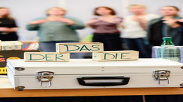 m Vordergrund stehen Holzklötze mit den deutschen Artikeln „der“, „das“ und „die“; im unscharfen Hintergrund sind mehrere Personen in einem Gruppenraum zu sehen.