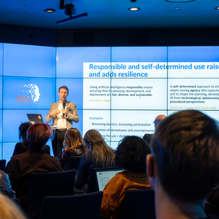 Ein Mann hält einen Vortrag vor Publikum mit einer Präsentation zum Thema verantwortungsvolle und selbstbestimmte Nutzung von Künstlicher Intelligenz auf mehreren Bildschirmen im Hintergrund.