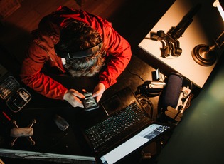 Perspektive von oben: Person, Content Creator, sitzt am Schreibtisch vor seinem Rechner und h&auml;lt sein Handy in der Hand und hat Kopfh&ouml;rer auf