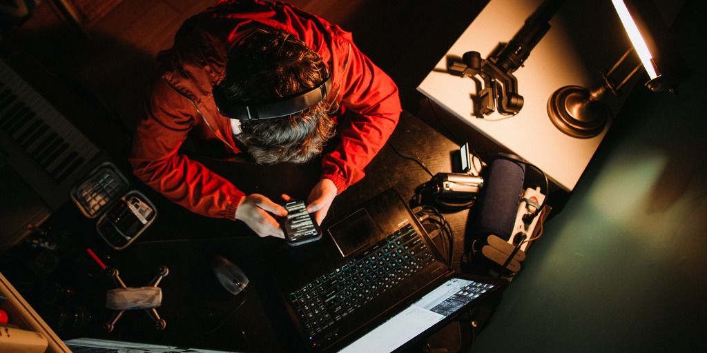 Perspektive von oben auf den Kopf von einem Mann mit Kopfhöhrern der auf sein Handybildschirm schaut. Er sitzt an einem Schreibtisch und sein Laptop ist aufgeklappt und angeschaltet. Neben dem Laptop sieht man außerdem noch eine Videokamera sowie ein kleines Stativ liegen.