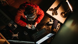 Perspektive von oben auf den Kopf von einem Mann mit Kopfhöhrern der auf sein Handybildschirm schaut. Er sitzt an einem Schreibtisch und sein Laptop ist aufgeklappt und angeschaltet. Neben dem Laptop sieht man außerdem noch eine Videokamera sowie ein kleines Stativ liegen.