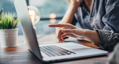 Drei Personen sitzen an einem Tisch. Eine Person zeigt auf den Bildschirm eines aufgeklappten Laptops. Auf der anderen Seite sitzt die dritte Person und hat einen aufgeklappten Laptop vor sich stehen. Im Hintergrund steht eine Pflanze. Die Sicht geht durch ein Fenster. Sie ist unscharf. 