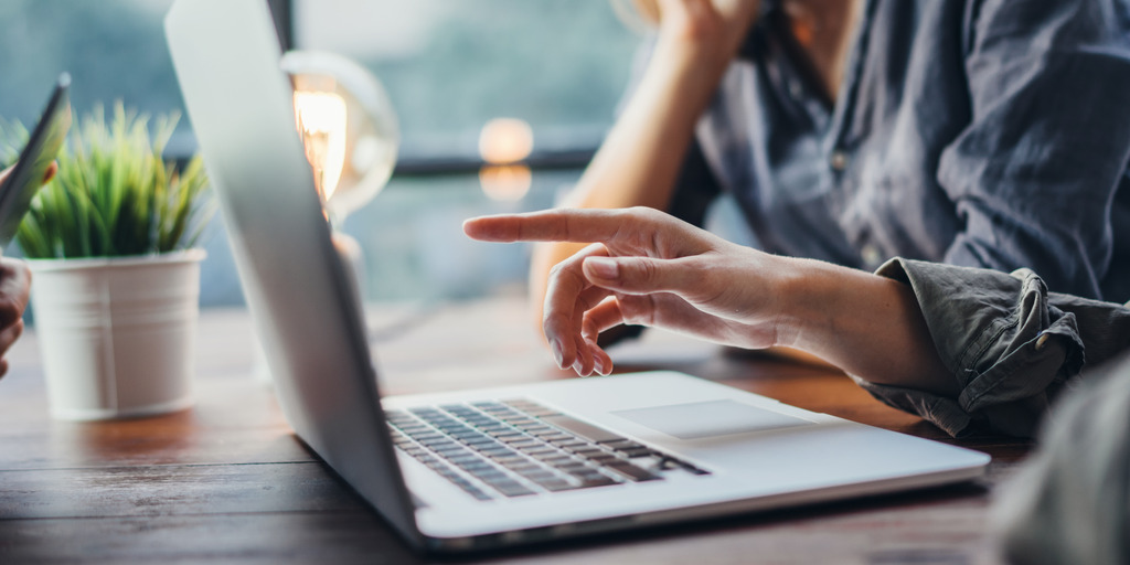 Drei Personen sitzen an einem Tisch. Eine Person zeigt auf den Bildschirm eines aufgeklappten Laptops. Auf der anderen Seite sitzt die dritte Person und hat einen aufgeklappten Laptop vor sich stehen. Im Hintergrund steht eine Pflanze. Die Sicht geht durch ein Fenster. Sie ist unscharf. 