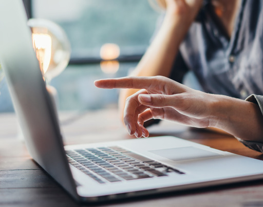 Drei Personen sitzen an einem Tisch. Eine Person zeigt auf den Bildschirm eines aufgeklappten Laptops. Auf der anderen Seite sitzt die dritte Person und hat einen aufgeklappten Laptop vor sich stehen. Im Hintergrund steht eine Pflanze. Die Sicht geht durch ein Fenster. Sie ist unscharf.