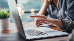 Drei Personen sitzen an einem Tisch. Eine Person zeigt auf den Bildschirm eines aufgeklappten Laptops. Auf der anderen Seite sitzt die dritte Person und hat einen aufgeklappten Laptop vor sich stehen. Im Hintergrund steht eine Pflanze. Die Sicht geht durch ein Fenster. Sie ist unscharf. 