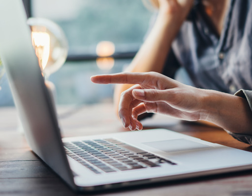 Drei Personen sitzen an einem Tisch. Eine Person zeigt auf den Bildschirm eines aufgeklappten Laptops. Auf der anderen Seite sitzt die dritte Person und hat einen aufgeklappten Laptop vor sich stehen. Im Hintergrund steht eine Pflanze. Die Sicht geht durch ein Fenster. Sie ist unscharf.
