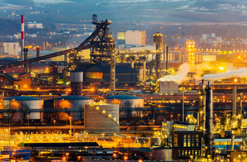 Industrieanlage bei D&auml;mmerung mit beleuchteten Fabrikhallen, Schornsteinen, Tanks und aufsteigendem Dampf.