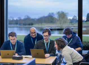 Das Foto zeigt eine kleine Gruppe von f&uuml;nf Personen in einem Workshop-/Seminarraum, die gemeinsam an Laptops arbeitet. Zwei sitzen am Tisch vor ihren Ger&auml;ten, drei weitere beugen sich dazu und schauen mit auf die Bildschirme.