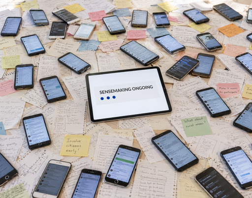 Zahlreiche Smartphones und bunte Zettel mit handschriftlichen Notizen liegen verstreut auf einer Fläche. In der Mitte befindet sich ein Tablet, auf dessen Bildschirm der englische Text „SENSEMAKING ONGOING“ mit drei blauen Punkten zu sehen ist. Dieses Bild wurde mit der KI generiert.