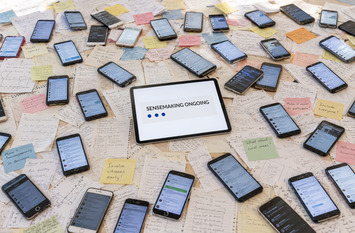 Ein Tisch ist komplett mit Notizzetteln und Smartphones bedeckt. In der Mitte liegt ein Tablet, auf dem die Statusanzeige "Sensemaking Ongoing" (Sinnbestimmung l&auml;uft) zu sehen ist.