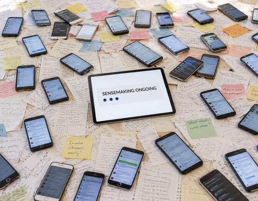 Zahlreiche Smartphones und bunte Zettel mit handschriftlichen Notizen liegen verstreut auf einer Fläche. In der Mitte befindet sich ein Tablet, auf dessen Bildschirm der englische Text „SENSEMAKING ONGOING“ mit drei blauen Punkten zu sehen ist. Dieses Bild wurde mit der KI generiert.