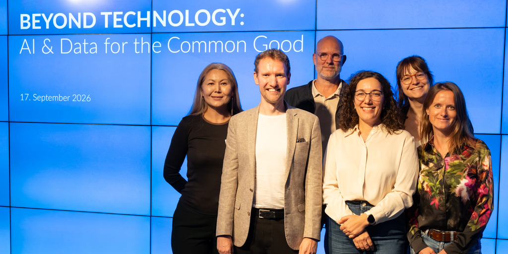 Sechs Personen stehen vor einer blauen Wand mit der Aufschrift 'BEYOND TECHNOLOGY: AI & Data for the Common Good 17. September 2026' und lächeln in die Kamera