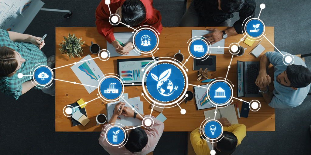 Eine Gruppe von Menschen sitzt an einem Holztisch in einer Besprechung, darüber liegt eine digitale Grafik vernetzter Icons zu den Themen Nachhaltigkeit, Umweltschutz und globale Vernetzung.