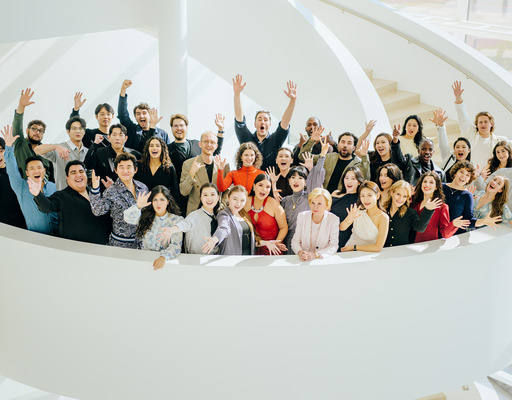 Gruppenfoto der Teilnehmer:innen der NEUEN STIMMEN 2026 zusammen mit Liz Mohn, Präsidentin der NEUEN STIMMEN, auf einer Wendeltreppe im Theater Gütersloh. Die Sänger:innen winken in die Kamera.