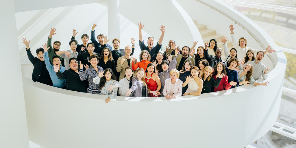 Gruppenfoto der Teilnehmer:innen der NEUEN STIMMEN 2026 zusammen mit Liz Mohn, Präsidentin der NEUEN STIMMEN, auf einer Wendeltreppe im Theater Gütersloh. Die Sänger:innen winken in die Kamera.