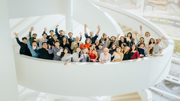 Gruppenfoto der Teilnehmer:innen der NEUEN STIMMEN 2026 zusammen mit Liz Mohn, Präsidentin der NEUEN STIMMEN, auf einer Wendeltreppe im Theater Gütersloh. Die Sänger:innen winken in die Kamera.