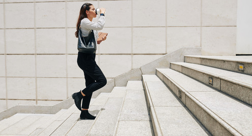 Junge Frau geht mit einem Kaffeebecher in der Hand eine weiße Treppe hoch.