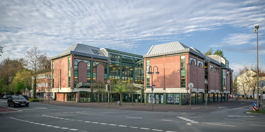 Außenansicht der Stadtbibliothek Gütersloh mit rosa Fassaden elements, grünen Fensterrahmen und einem verglasten Mittelbereich. Das mehrstöckige Gebäude steht an einer gepflasterten Kreuzung unter leicht bewölktem Himmel.