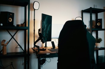 Arbeitsplatz von Creator:innen zu sehen mit Schreibtisch, Computer, B&uuml;rostuhl, Beleuchtung und einem Regal