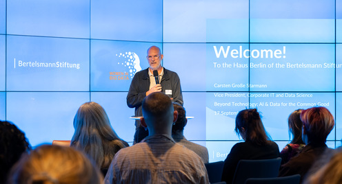 Ein Mann steht mit Mikrofon vor einem Publikum und spricht vor einer großen Bildschirmwand mit Text und Logos der Bertelsmann Stiftung und Women in Big Data