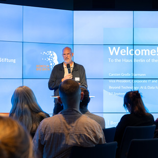 Ein Mann steht mit Mikrofon vor einem Publikum und spricht vor einer großen Bildschirmwand mit Text und Logos der Bertelsmann Stiftung und Women in Big Data