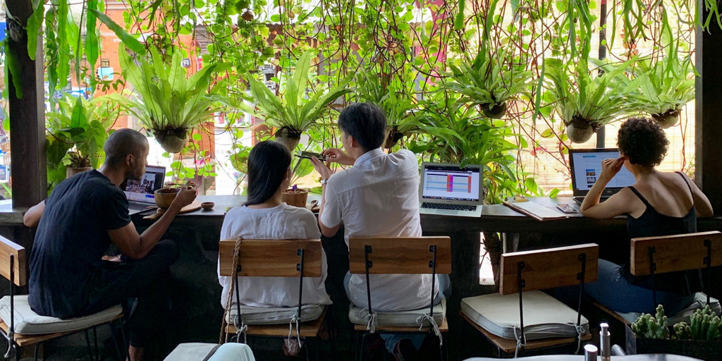 In einem Lokal auf der indonesischen Insel Bali sind mehrere junge Menschen von hinten zu sehen, wie sie an einem langen Tisch vor ihren Laptops sitzen und arbeiten oder essen.