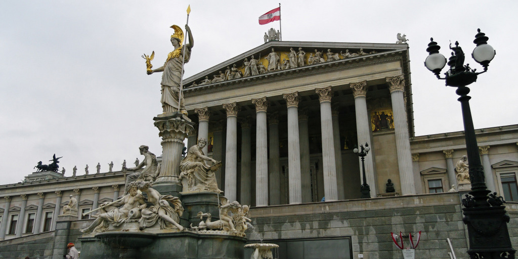 Blick auf den Nationalrat, das österreichische Parlamentsgebäude, in Wien. Davor ein Springbrunnen und eine Straßenlaterne.