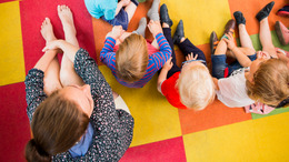 Mehrere Kinder sitzen in einem Sitzkreis auf einem bunten Teppich in der Kita "Blumenkinder" und die Erzieherin sitzt daneben - zu sehen aus der Vogelperspektive.