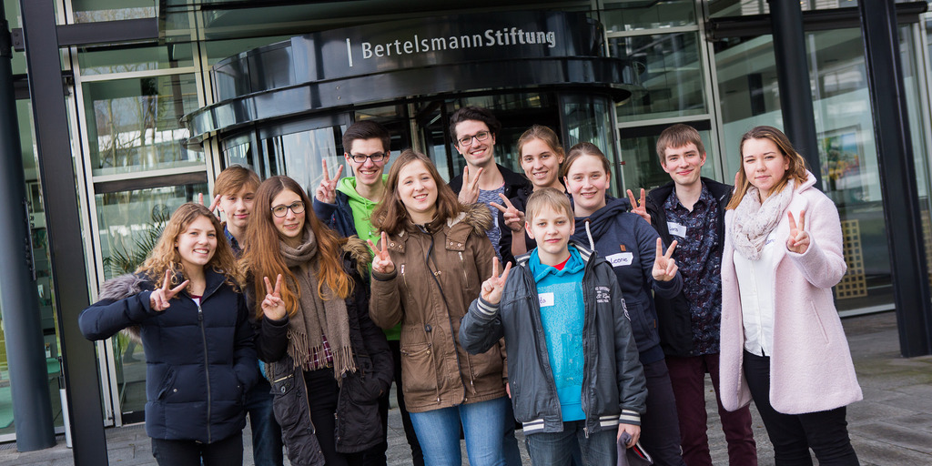 Die "Alle Kids sind VIPs"-Kinder Jury 2016 steht vor dem Eingang der Bertelsmann-Stiftung und zeigt das Peacezeichen. Es ist eine Gruppe von 11 Jugendlichen, die aus Jungen und Mädchen besteht.