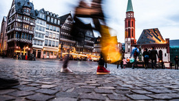 [Translate to English:] Blick auf einen Marktplatz einer Stadt mit vielen alten Gebäuden. Es sind verschwommene Umrisse im Bild zu sehen von einer Person die gerade vorbeigeht.